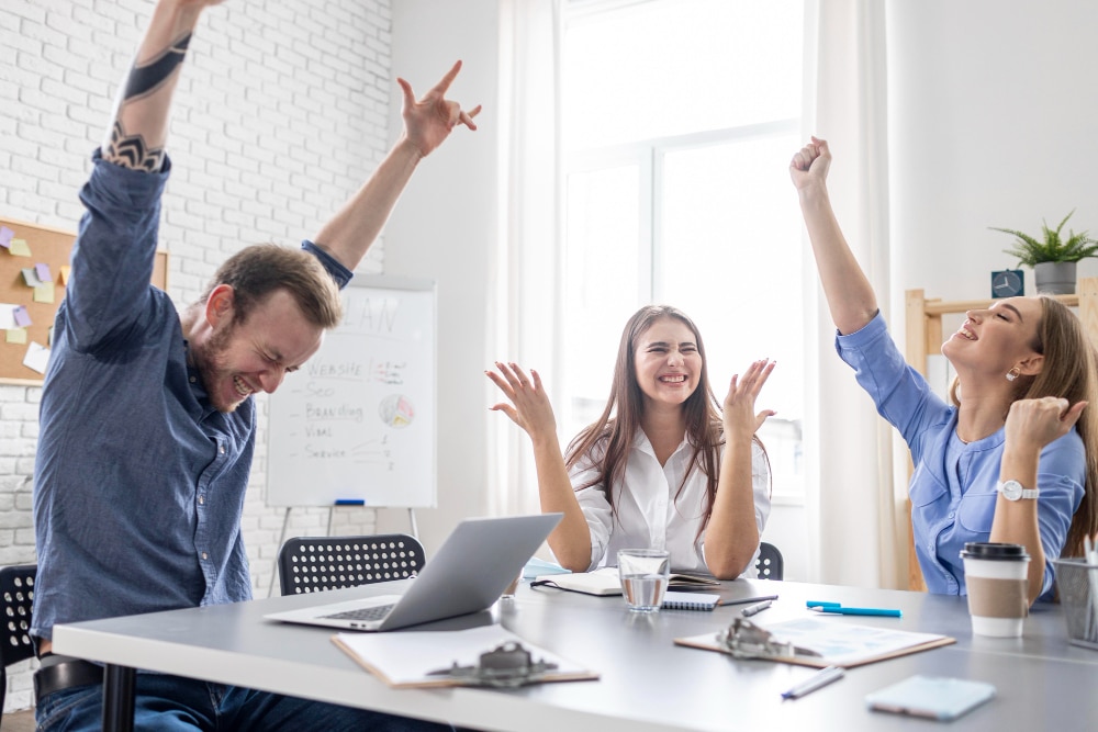3 personnes heureuses autour d'un bureau avec les bras levés pour illustrer les avantages les plus recherchés par les candidats en 2023.